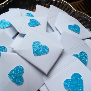 Peut inclure: Gros plan sur de petites enveloppes blanches, chacune orn&eacute;e d'un c&oelig;ur scintillant turquoise. Les enveloppes sont dispos&eacute;es dans un plat d&eacute;coratif &agrave; bord dor&eacute;, cr&eacute;ant une pr&eacute;sentation charmante et festive.