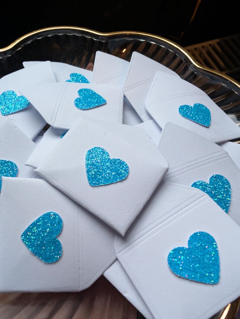 Peut inclure: Gros plan sur de petites enveloppes blanches, chacune orn&eacute;e d'un c&oelig;ur scintillant turquoise. Les enveloppes sont dispos&eacute;es dans un plat d&eacute;coratif &agrave; bord dor&eacute;, cr&eacute;ant une pr&eacute;sentation charmante et festive.