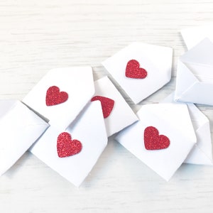 Peut inclure: Plusieurs petites enveloppes blanches en papier, chacune orn&eacute;e d'un c&oelig;ur rouge paillet&eacute;. Les enveloppes sont dispos&eacute;es sur une surface en bois clair, &eacute;voquant l'amour ou la Saint-Valentin.