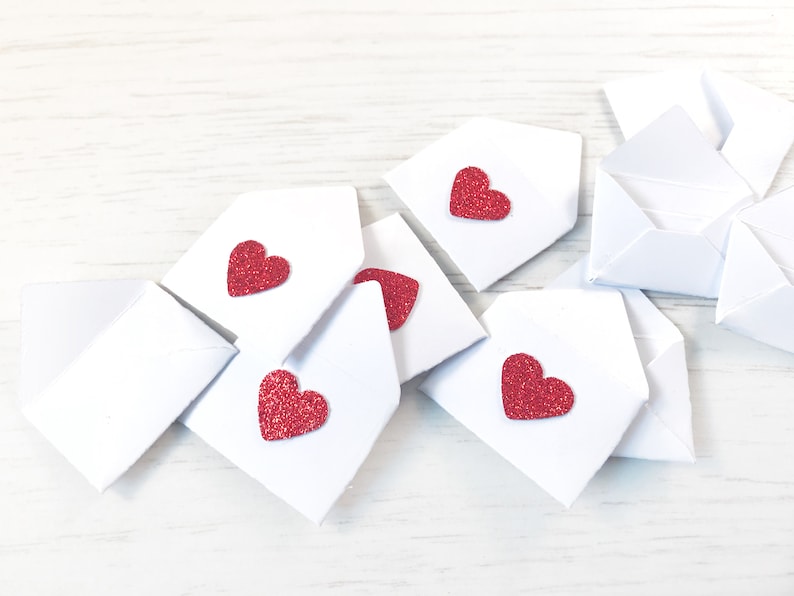 Peut inclure: Plusieurs petites enveloppes blanches en papier, chacune orn&eacute;e d'un c&oelig;ur rouge paillet&eacute;. Les enveloppes sont dispos&eacute;es sur une surface en bois clair, &eacute;voquant l'amour ou la Saint-Valentin.