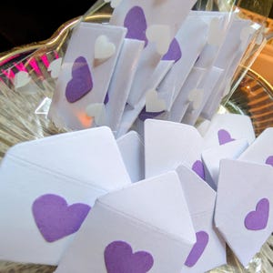 Peut inclure: Enveloppes en papier blanc avec des c&oelig;urs violets dispos&eacute;es dans un bol en verre transparent. Certaines enveloppes sont emball&eacute;es individuellement dans du plastique transparent. De petits autocollants en forme de c&oelig;ur blanc sont &eacute;galement visibles.