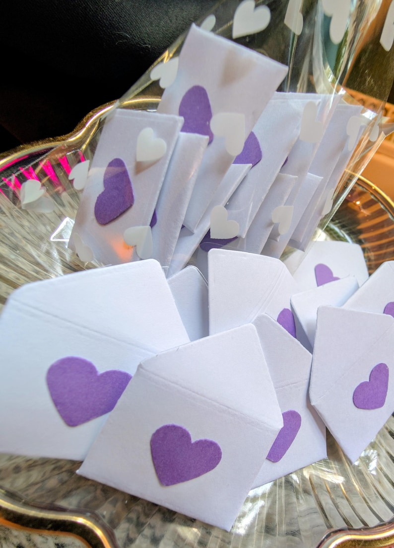 Peut inclure: Enveloppes en papier blanc avec des c&oelig;urs violets dispos&eacute;es dans un bol en verre transparent. Certaines enveloppes sont emball&eacute;es individuellement dans du plastique transparent. De petits autocollants en forme de c&oelig;ur blanc sont &eacute;galement visibles.