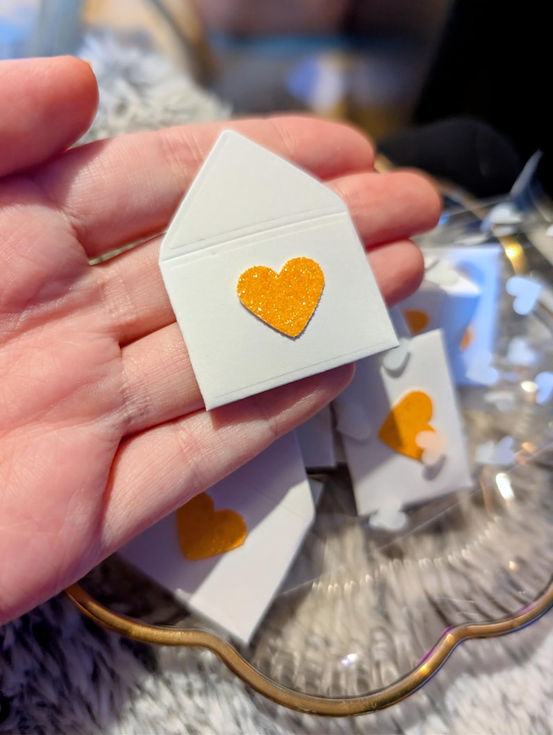 Peut inclure: Petites enveloppes en papier blanc avec un c&oelig;ur orange scintillant. Plusieurs enveloppes sont visibles, certaines nettes, d'autres floues. Les enveloppes sont dispos&eacute;es dans un plat d&eacute;coratif transparent.