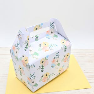 May include: A white gift box with a floral pattern and a handle. The box is decorated with a repeating design of watercolour flowers in pastel shades, including yellow, peach, and blue. It rests on a yellow surface.