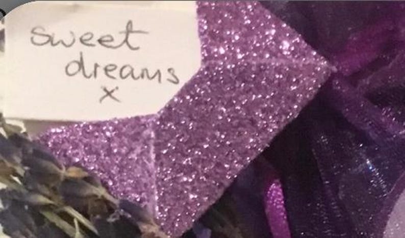 Peut inclure: Gros plan sur un objet d&eacute;coratif avec un n&oelig;ud violet scintillant et une petite &eacute;tiquette blanche. L'&eacute;tiquette porte les mots manuscrits "sweet dreams x". L'arri&egrave;re-plan comprend de la lavande s&eacute;ch&eacute;e et du tissu violet fonc&eacute;.