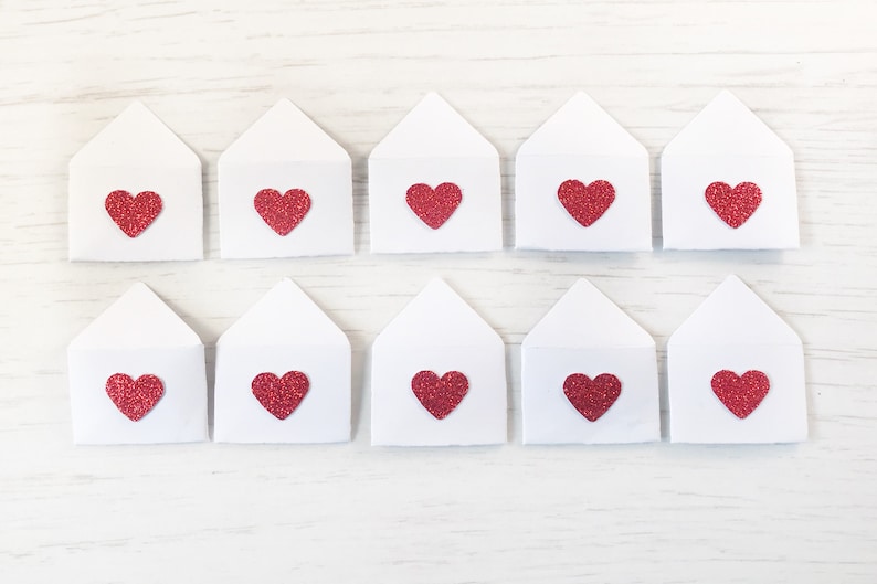 Peut inclure: Dix enveloppes en papier blanc, chacune orn&eacute;e d'un c&oelig;ur rouge paillet&eacute;. Les enveloppes sont dispos&eacute;es en deux rang&eacute;es de cinq sur une surface en bois blanc. Les c&oelig;urs sont d'un rouge vif.