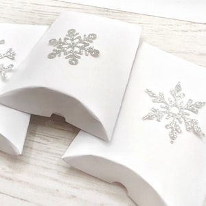 May include: Three white pillow boxes, each adorned with a silver glitter snowflake. The boxes are displayed on a light wooden surface, evoking a winter theme. Suitable for small gifts or festive favours.