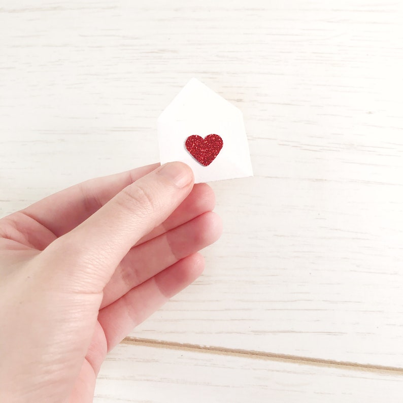 Peut inclure: Une petite enveloppe en papier blanc avec un c&oelig;ur rouge paillet&eacute; au centre. L'enveloppe est tenue dans une main sur un fond en bois blanc. Le c&oelig;ur est d'un rouge vif et scintillant.