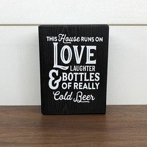May include: Black wooden sign with white text that reads "This house runs on love, laughter & bottles of really cold beer."