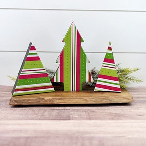 May include: Three wooden Christmas tree decorations with a red, white and green striped and polka dot pattern. The decorations are sitting on a wooden base.