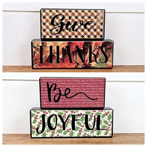 May include: A set of four wooden blocks with a fall-themed design. The blocks spell out "Give Thanks Be Joyful". The top block has a plaid pattern in shades of brown, yellow, and red. The second block has a red and orange background with a pattern of autumn leaves. The third block has a red background with a pattern of the words "Merry Christmas". The bottom block has a red background with a pattern of holly berries and leaves.