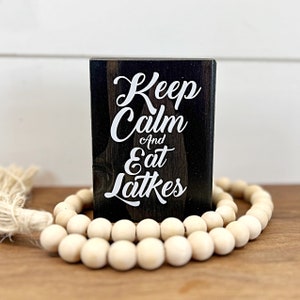May include: A black wooden sign with white text that reads "Keep Calm and Eat Latkes". The sign is sitting on a table with a string of white wooden beads.