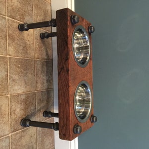 May include: A rustic, wall-mounted pet feeder with two stainless steel bowls set into a reclaimed wood frame. The frame is attached to the wall with black metal pipes and fittings. The wood has a reddish-brown hue.