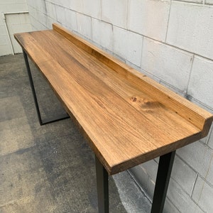 May include: A long, wooden table with a natural wood finish and black metal legs. The table top has a slightly raised edge.