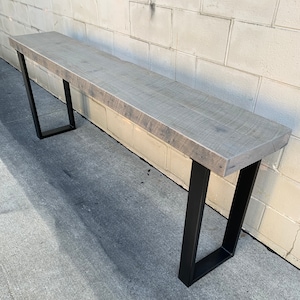 May include: A long, rustic wooden console table with a gray stain and black metal legs. The table is sitting on a concrete surface.