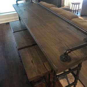 May include: A wooden bar table with black metal pipe legs and a black metal pipe handrail. The table has a rustic wood finish and is accompanied by three matching bar stools.
