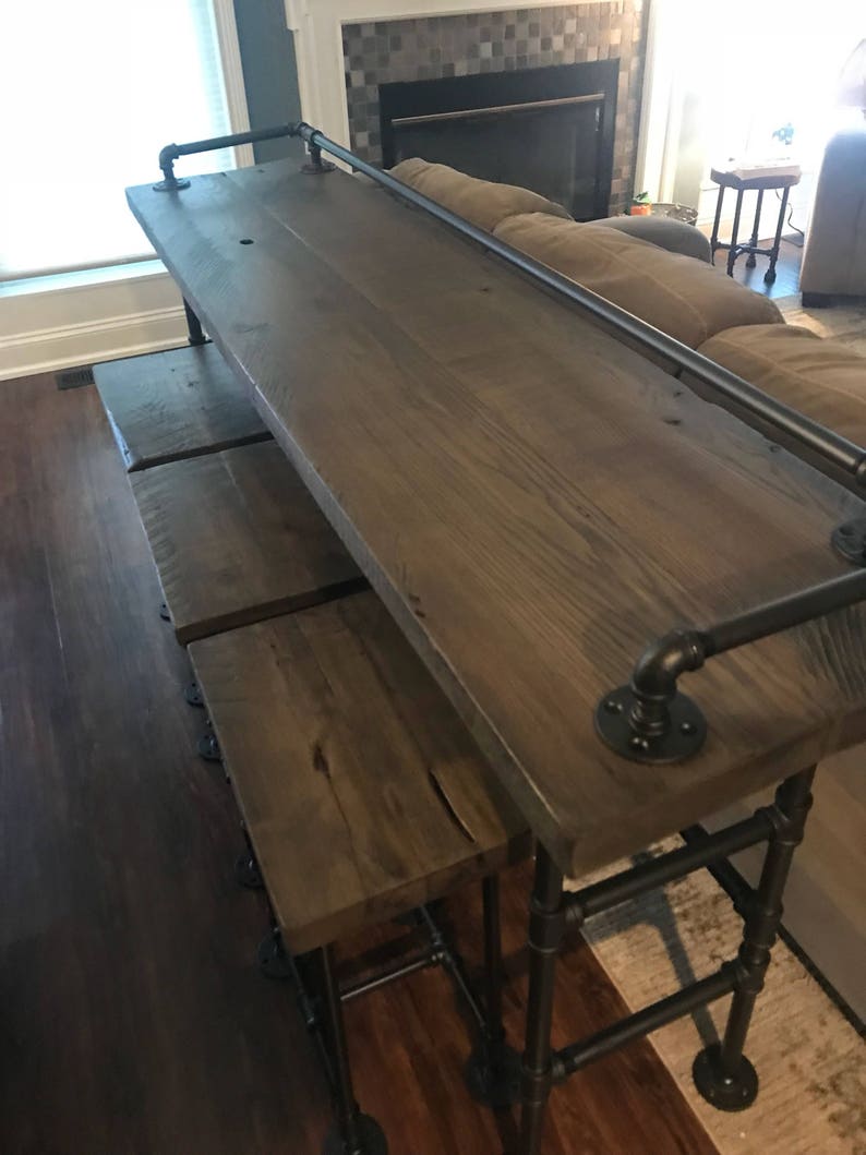 May include: A wooden bar table with black metal pipe legs and a black metal pipe handrail. The table has a rustic wood finish and is accompanied by three matching bar stools.