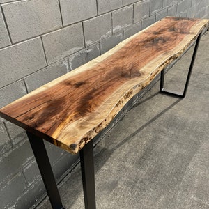 May include: A long, live edge walnut wood table with a clear epoxy finish. The table has a natural, rustic look and is supported by two black metal legs.