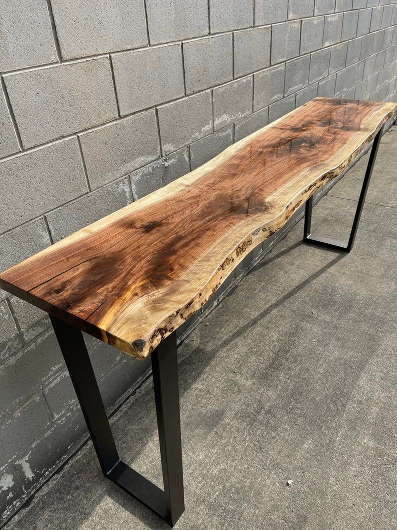 May include: A long, live edge walnut wood table with a clear epoxy finish. The table has a natural, rustic look and is supported by two black metal legs.