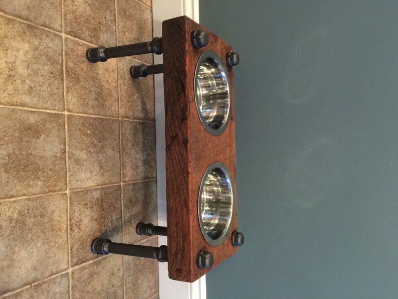 May include: A rustic, wall-mounted pet feeder with two stainless steel bowls set into a reclaimed wood frame. The frame is attached to the wall with black metal pipes and fittings. The wood has a reddish-brown hue.