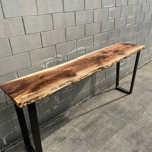 May include: A long, live edge walnut wood table with a natural, light brown finish. The table has a black metal frame with a simple, rectangular design. The table is sitting on a concrete surface.