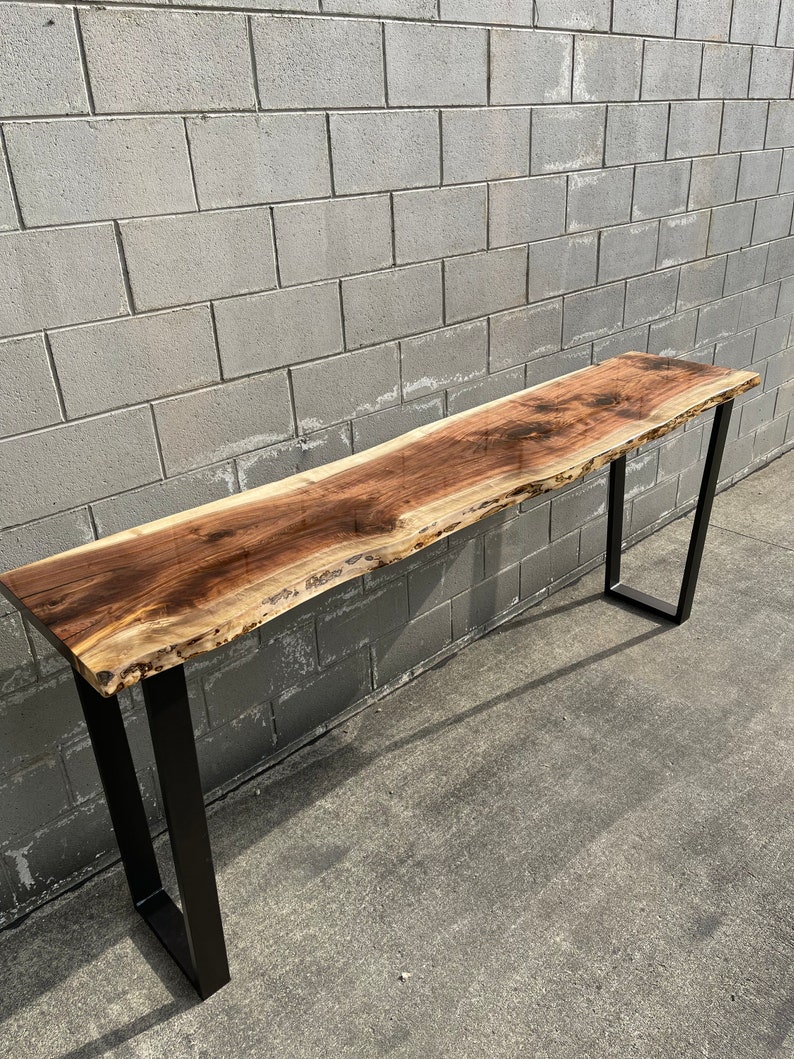 May include: A long, live edge walnut wood table with a natural, light brown finish. The table has a black metal frame with a simple, rectangular design. The table is sitting on a concrete surface.