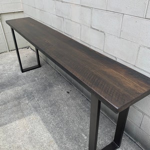 May include: A long, dark brown wooden console table with a black metal frame. The table has a rustic, reclaimed wood look and is supported by two rectangular metal legs.