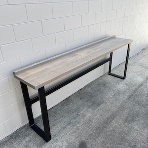 May include: A long, rectangular table with a light gray wood top and a black metal frame. The table is standing on a gray concrete floor.