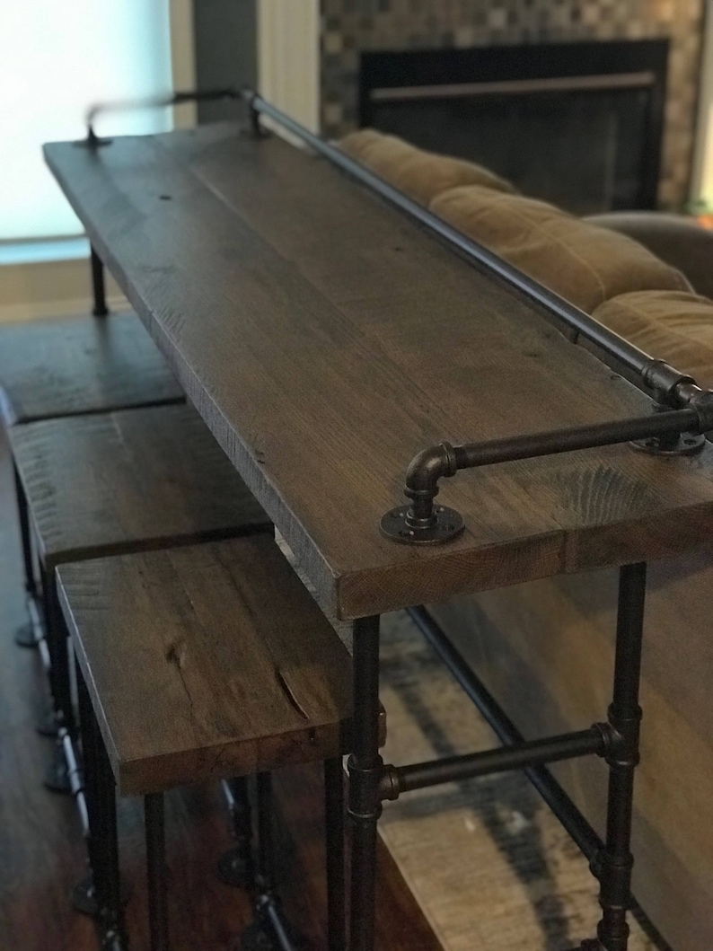 May include: A rustic wooden bar table with black metal pipe legs and a top shelf. The table is paired with two matching wooden stools with black metal pipe legs.