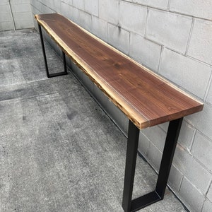 May include: A long, live edge walnut wood table with a natural, rustic finish. The table is supported by two black metal legs.