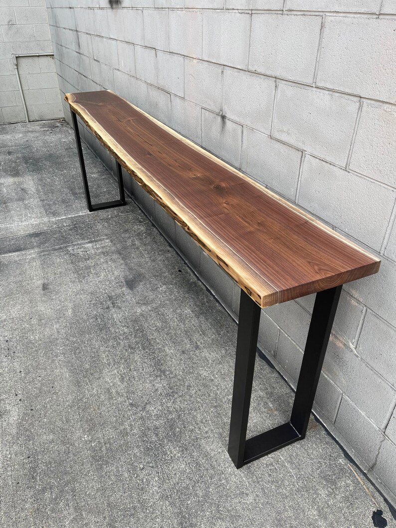 May include: A long, live edge walnut wood table with a natural, rustic finish. The table is supported by two black metal legs.