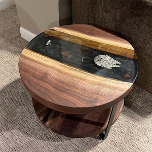 May include: A round wooden end table with a black epoxy resin top. The top has a clear coat and a small silver spaceship model embedded in the resin. The table has a lower shelf made of wood.