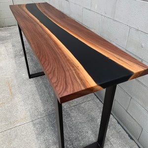 May include: A long, narrow walnut wood table with a black epoxy river running down the center. The table is supported by two black metal legs.