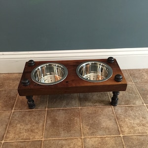 May include: A raised dog bowl stand with two stainless steel bowls set into a dark stained wooden frame. The stand has black pipe legs and black accents on the corners. The bowls are designed to hold food and water for pets.