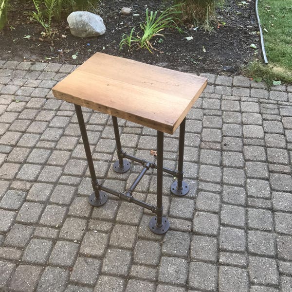 May include: A wooden table top with a rustic finish sits on a black metal pipe base. The table is on a gray brick patio.