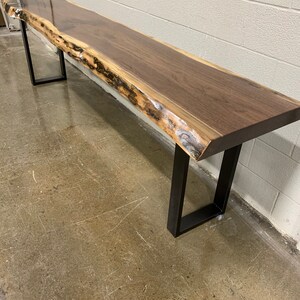 May include: A long, dark brown wooden bench with a live edge top and black metal legs. The bench is sitting on a concrete floor.