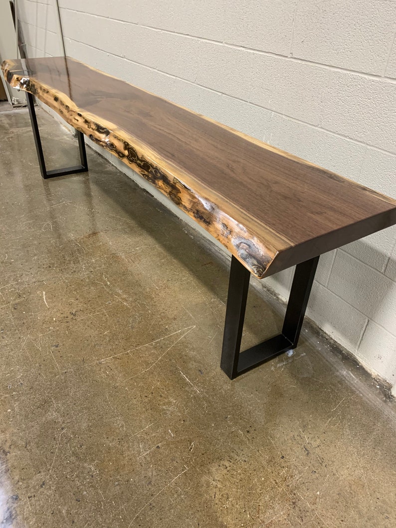 May include: A long, dark brown wooden bench with a live edge top and black metal legs. The bench is sitting on a concrete floor.