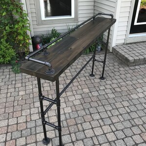 May include: A black metal bar table with a dark wood top and a black metal handrail. The table is standing on a gray brick patio.