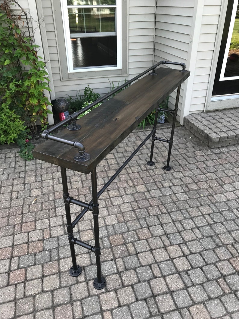 May include: A black metal bar table with a dark wood top and a black metal handrail. The table is standing on a gray brick patio.