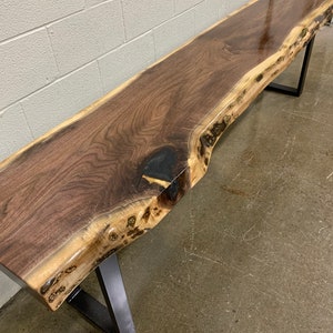 May include: A long, dark brown wooden bench with a live edge top and black metal legs. The bench is made of walnut wood and has a natural, rustic look.
