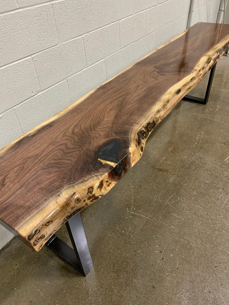 May include: A long, dark brown wooden bench with a live edge top and black metal legs. The bench is made of walnut wood and has a natural, rustic look.