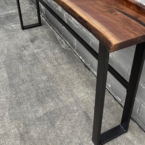 May include: A long, dark brown wooden console table with a live edge top and black metal legs. The table is standing on a concrete floor.