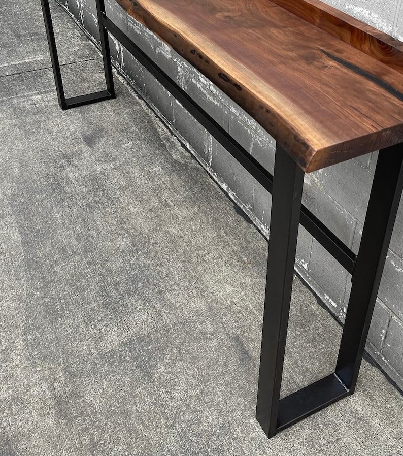 May include: A long, dark brown wooden console table with a live edge top and black metal legs. The table is standing on a concrete floor.