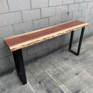 May include: A long, narrow wooden table with a live edge top and black metal legs. The table is made of wood with a natural grain pattern and a reddish-brown color.