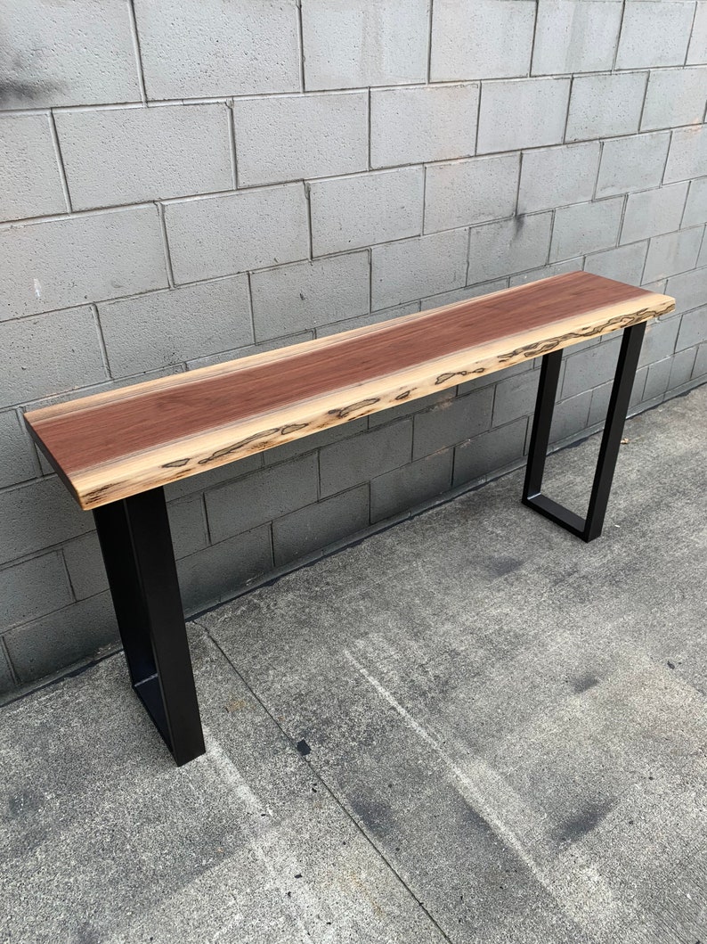 May include: A long, narrow wooden table with a live edge top and black metal legs. The table is made of wood with a natural grain pattern and a reddish-brown color.