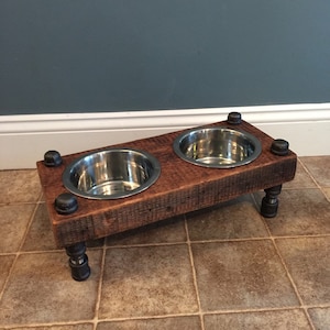 May include: A rustic, elevated pet feeder with two stainless steel bowls set into a reclaimed wood frame. The frame is supported by black pipe fittings, creating a sturdy and stylish design. The feeder measures approximately 24 inches long.