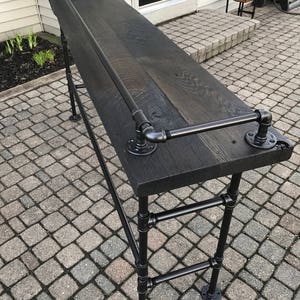 May include: A black wooden bar table with a black metal pipe frame. The table has a black metal pipe handrail and a black metal pipe shelf. The table is sitting on a brick patio.