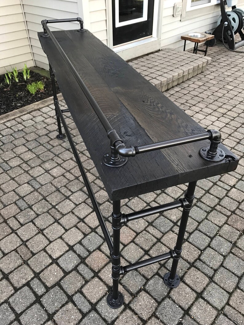 May include: A black wooden bar table with a black metal pipe frame. The table has a black metal pipe handrail and a black metal pipe shelf. The table is sitting on a brick patio.