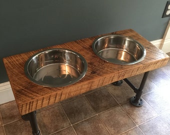 May include: A rustic, two-bowl pet feeder. The feeder features two stainless steel bowls set into a reclaimed wood surface. The wood is supported by black metal pipe legs. The feeder is designed to elevate the bowls.