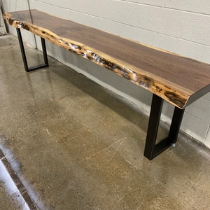 May include: A long, live edge walnut wood table with a natural, live edge finish. The table is supported by two black metal legs.